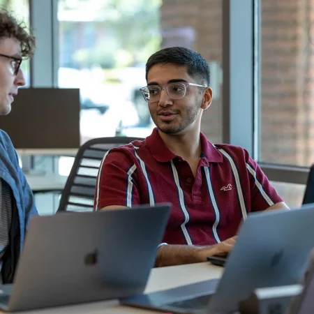 Dos estudiantes universitarios dialogan mientras trabajan con laptops en un aula.